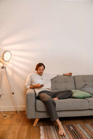 Ginger bristle man using mobile phone while resting on couch at homeの写真素材