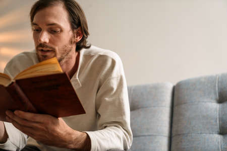 Ginger bristle man reading book while sitting on couch at home
