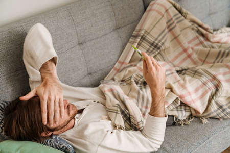 European man with flu measuring temperature while lying on sofa at homeの写真素材