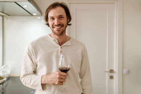 Ginger bristle man smiling while drinking wine in kitchen at homeの写真素材