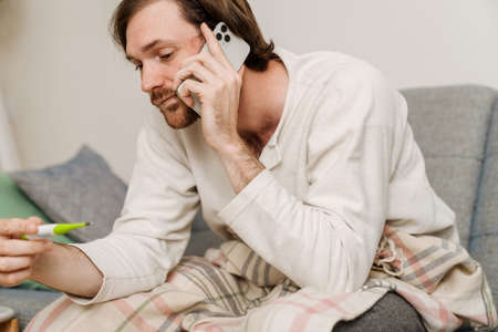 European man with flu talking on cellphone and measuring temperature at homeの写真素材