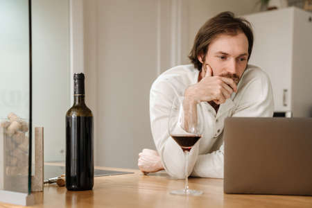 Ginger bristle man using laptop while drinking wine in kitchen at homeの写真素材