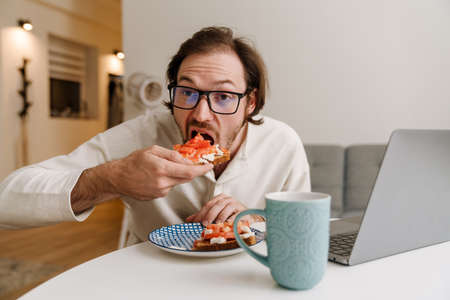 Ginger bristle man in eyeglasses working with laptop while having dinner at homeの写真素材