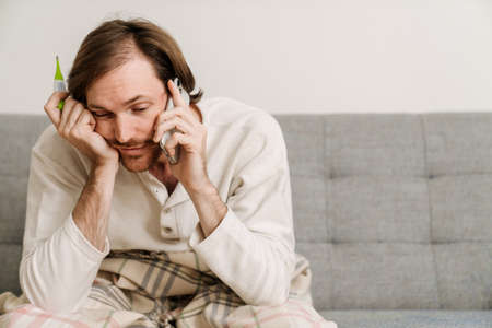 European man with flu talking on cellphone while sitting on sofa at homeの写真素材