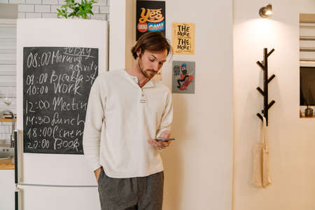 European unshaven man using mobile phone while standing in kitchen at home