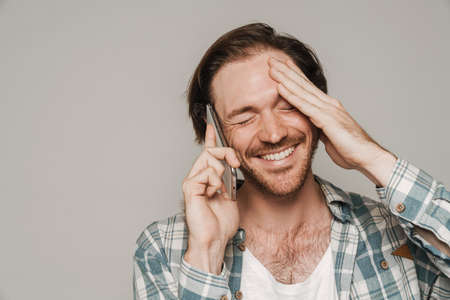 Bristle man dressed in shirt laughing while talking on cellphone isolated over grey background