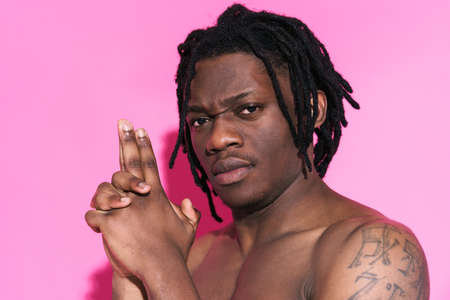 Black shirtless man with dreadlocks frowning while making gun gesture isolated over pink backgroundの写真素材