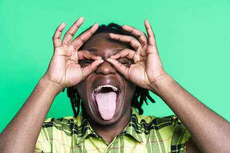 Young black man with dreadlocks gesturing and showing her tongue isolated over green backgroundの写真素材