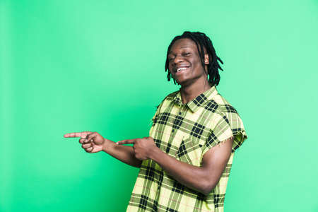 Young black man with dreadlocks smiling and pointing fingers aside isolated over green background