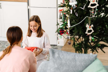 White mother giving her happy daughter present by christmas tree at homeの写真素材