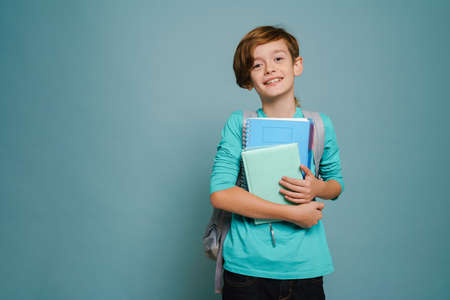 Ginger white boy smiling while posing with exercise books isolated over blue wallの写真素材