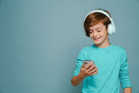 Ginger boy smiling while listening music with headphones and cellphone isolated over blue wallの写真素材