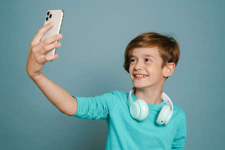 Ginger white boy with headphones taking selfie photo on cellphone isolated over blue wallの写真素材