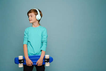Ginger white boy in headphones posing with skateboard isolated over blue wallの写真素材