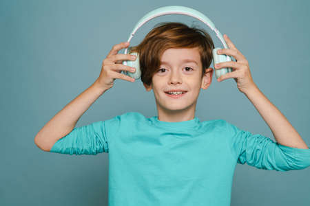 Ginger white boy smiling while listening music with headphones isolated over blue wallの写真素材