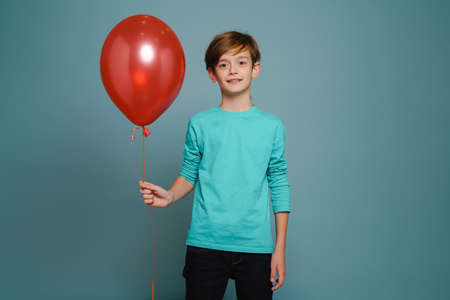 Ginger white boy smiling while posing with red balloon isolated over blue wallの写真素材