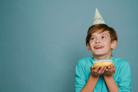 Ginger boy wearing party cone smiling while posing with doughnut isolated over blue wallの写真素材