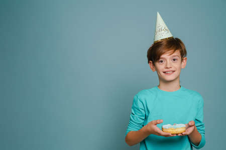 Ginger boy wearing party cone smiling while posing with doughnut isolated over blue wallの写真素材