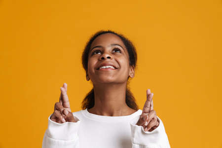 Brunette black girl smiling while holding fingers crossed for good luck isolated over yellow...