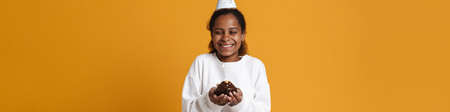 Brunette black girl in party cone posing with birthday cake isolated over yellow background