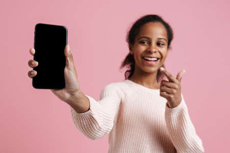 Black girl pointing finger and showing cellphone at camera isolated over pink backgroundの写真素材