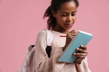 Black girl with backpack writing down notes in exercise book isolated over pink backgroundの写真素材