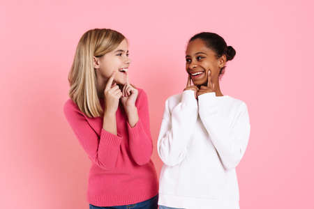 Multiracial two girls wearing sweaters pointing fingers at their smiles isolated over pink wallの写真素材