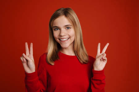 Blonde white girl laughing while showing peace sign isolated over red wallの写真素材