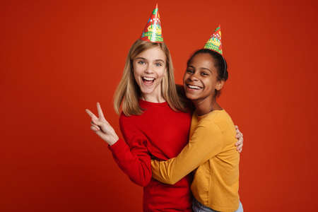 Multiracial two girls wearing party cones gesturing while hugging together isolated over red wallの写真素材