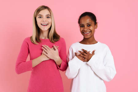 Multiracial two girls smiling while holding hands on their chests isolated over pink wallの写真素材