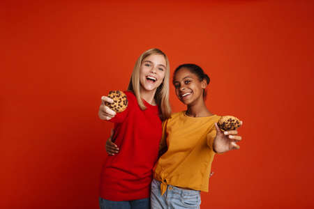 Multiracial two girls hugging while making fun with cookies isolated over red wallの写真素材