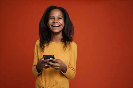 Brunette black girl smiling and using mobile phone isolated over red wall
