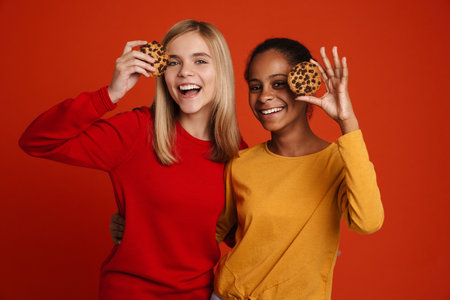 Multiracial two girls hugging while making fun with cookies isolated over red wall