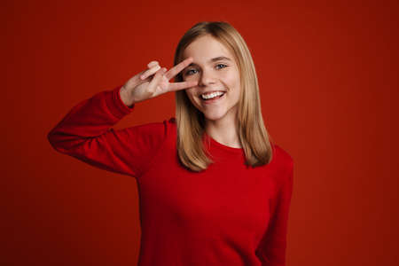 Blonde white girl laughing while showing peace sign isolated over red wall