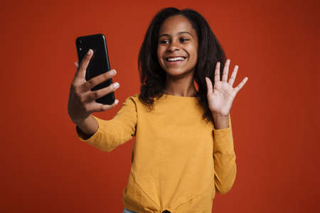 Brunette black girl waving hand and using mobile phone isolated over red wallの写真素材