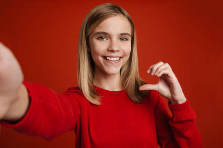 Blonde white girl gesturing while taking selfie photo isolated over red wall