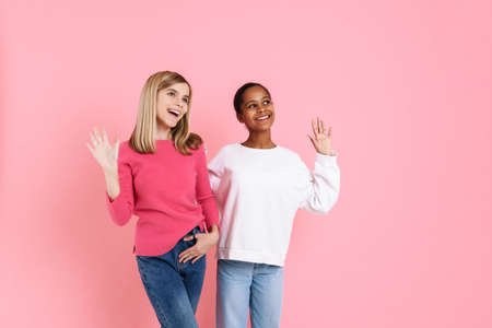 Multiracial two girls wearing sweaters smiling and waving hands together isolated over pink wallの写真素材