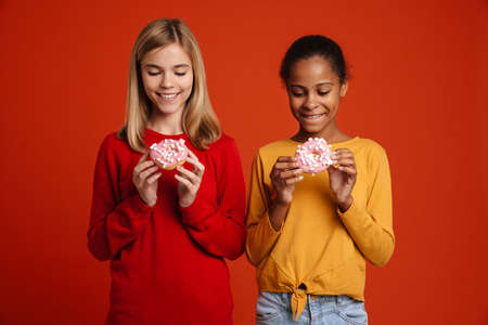 Multiracial two girls smiling while posing with donuts isolated over red wallの写真素材