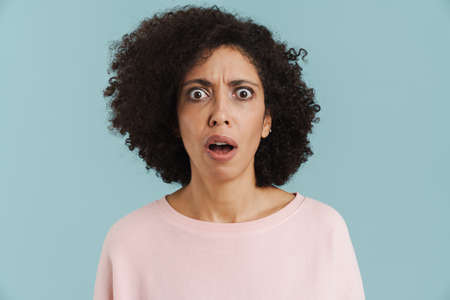 Young black woman with curly hair expressing surprise at camera isolated over blue background
