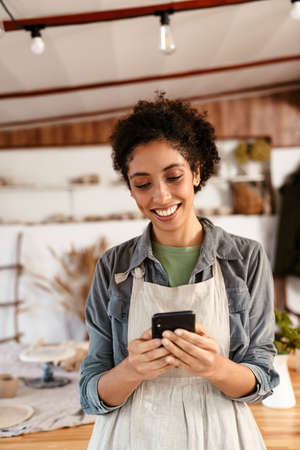 Young black ceramist woman wearing apron using cellphone while working in her studioの写真素材