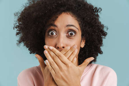 Young black woman covering her mouth while expressing surprise isolated over blue background