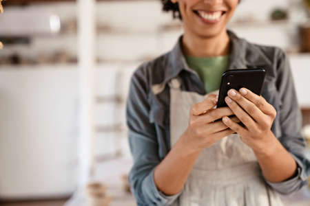 Young black ceramist woman wearing apron using cellphone while working in her studioの写真素材
