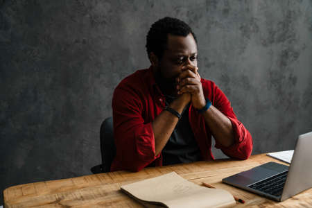 Black bearded man working with laptop while sitting at desk indoors