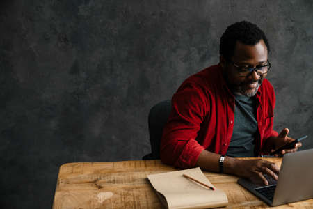 Black man in eyeglasses using cellphone while working with laptop indoorsの写真素材