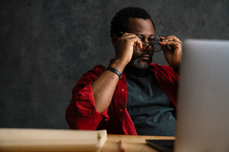 Black man in eyeglasses working with laptop while sitting at desk indoorsの写真素材