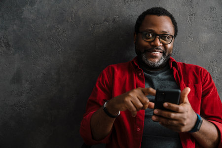 Black man in eyeglasses using cellphone while leaning on concrete wall indoorsの写真素材