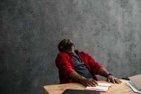 Black tired man in eyeglasses working with laptop while sitting at desk indoors