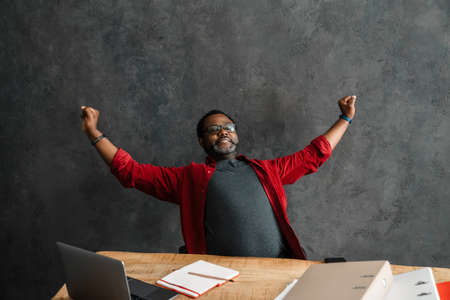 Black man in eyeglasses stretching his body while working with laptop indoorsの写真素材