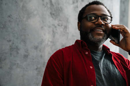 Black man in eyeglasses talking on cellphone while standing by concrete wall