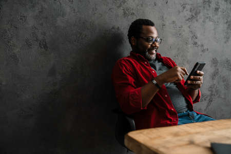 Black man in eyeglasses using cellphone while working at desk indoorsの写真素材